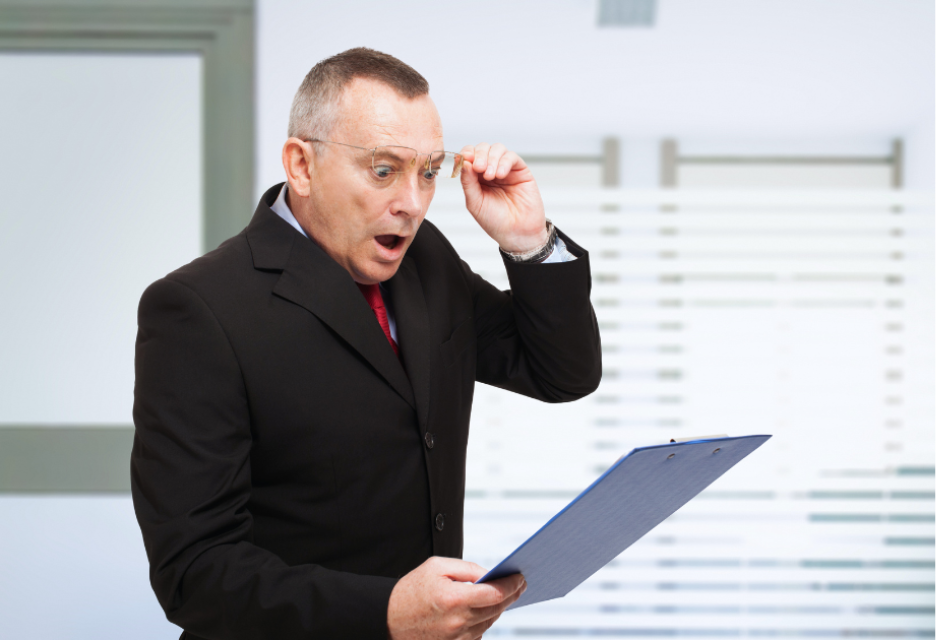 Surprised businessman in a black suit and red tie examines a document on a clipboard, reacting with shock to unexpected costs or budget overruns.